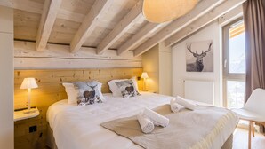 Teto de madeira, cama com lençóis brancos, cabeceira com estampas de veados, luminária, janela com cortinas e uma cadeira.