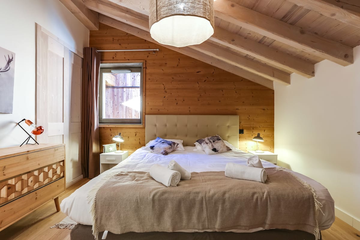 Une chambre confortable avec un plafond en bois, un grand lit recouvert d’une couverture beige, deux oreillers et une table de chevet avec une lampe.