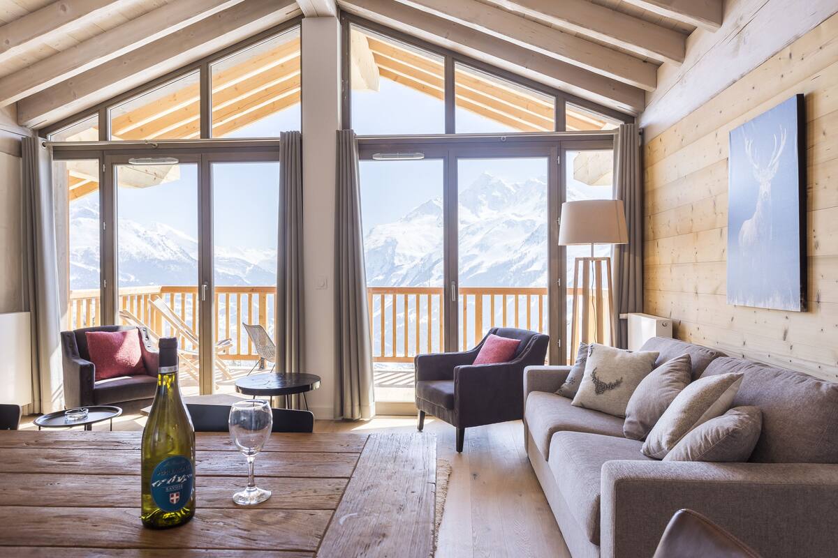 Une pièce chaleureuse avec un plafond en bois, de grandes fenêtres, une terrasse en bois, un canapé, un fauteuil, une table, une bouteille de vin et un verre.