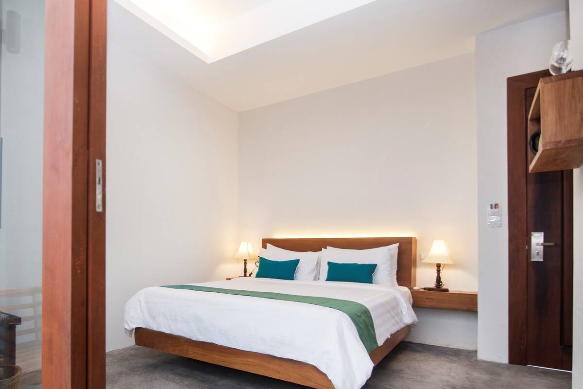Une chambre d’hôtel avec un lit, des lampes de chevet, une tête de lit en bois et une petite étagère en bois.