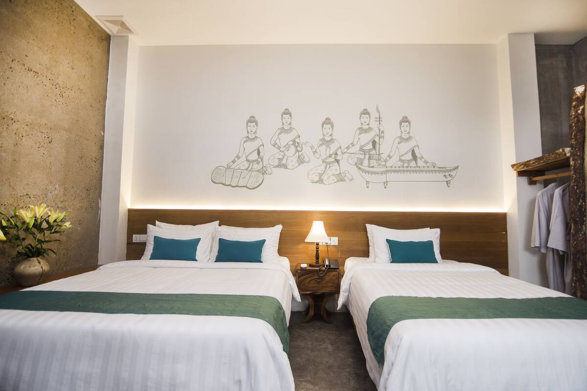 Une chambre d’hôtel avec deux lits, une table de chevet avec une lampe, une œuvre d’art murale représentant des personnages vêtus de costumes traditionnels, et un vase avec des fleurs sur le côté gauche.
