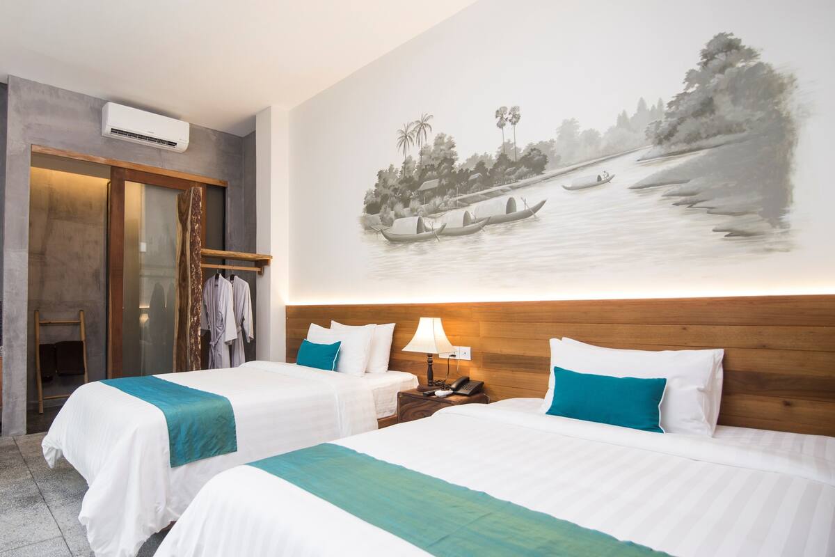 Une chambre d’hôtel avec deux lits, une tête de lit en bois, un climatiseur fixé au mur, un tableau représentant un paysage fluvial et une petite table avec une lampe.