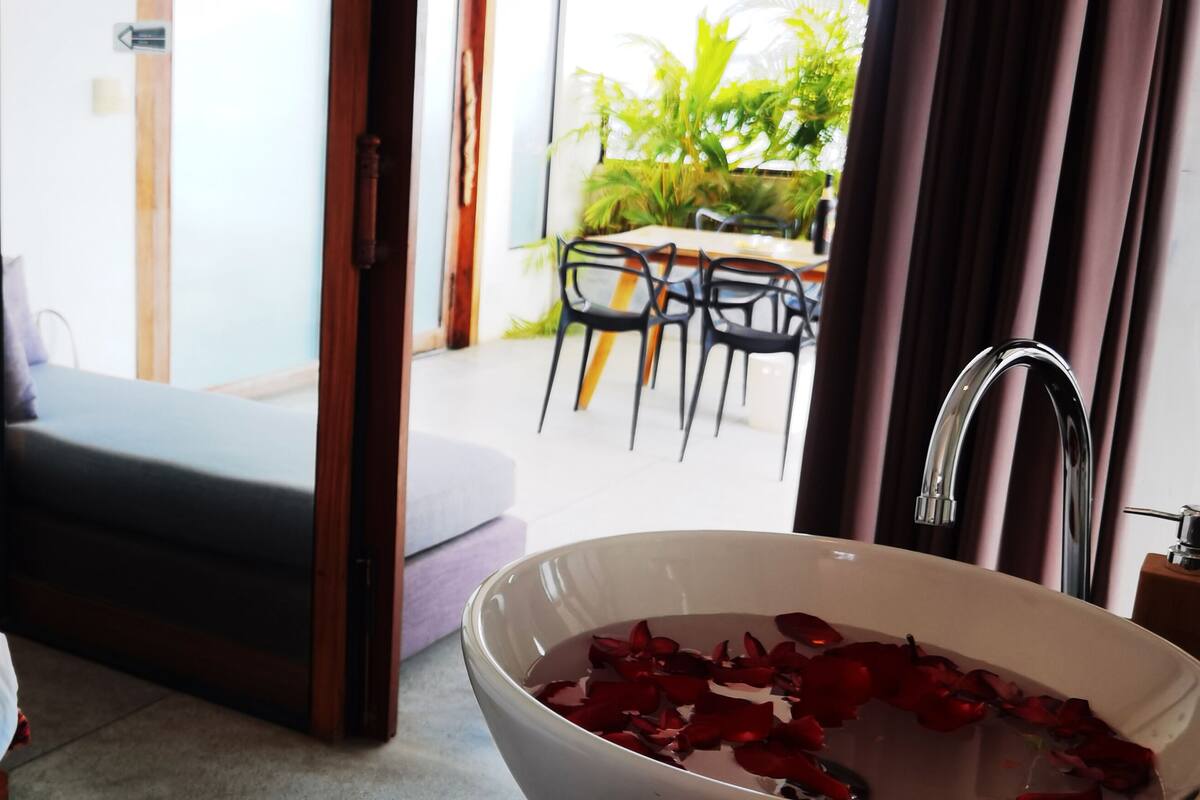 Une chambre d’hôtel avec un lit, une salle de bain dotée d’un grand lavabo rempli d’eau et de pétales de rose, un coin repas avec des chaises et une table, et une vue sur l’extérieur.