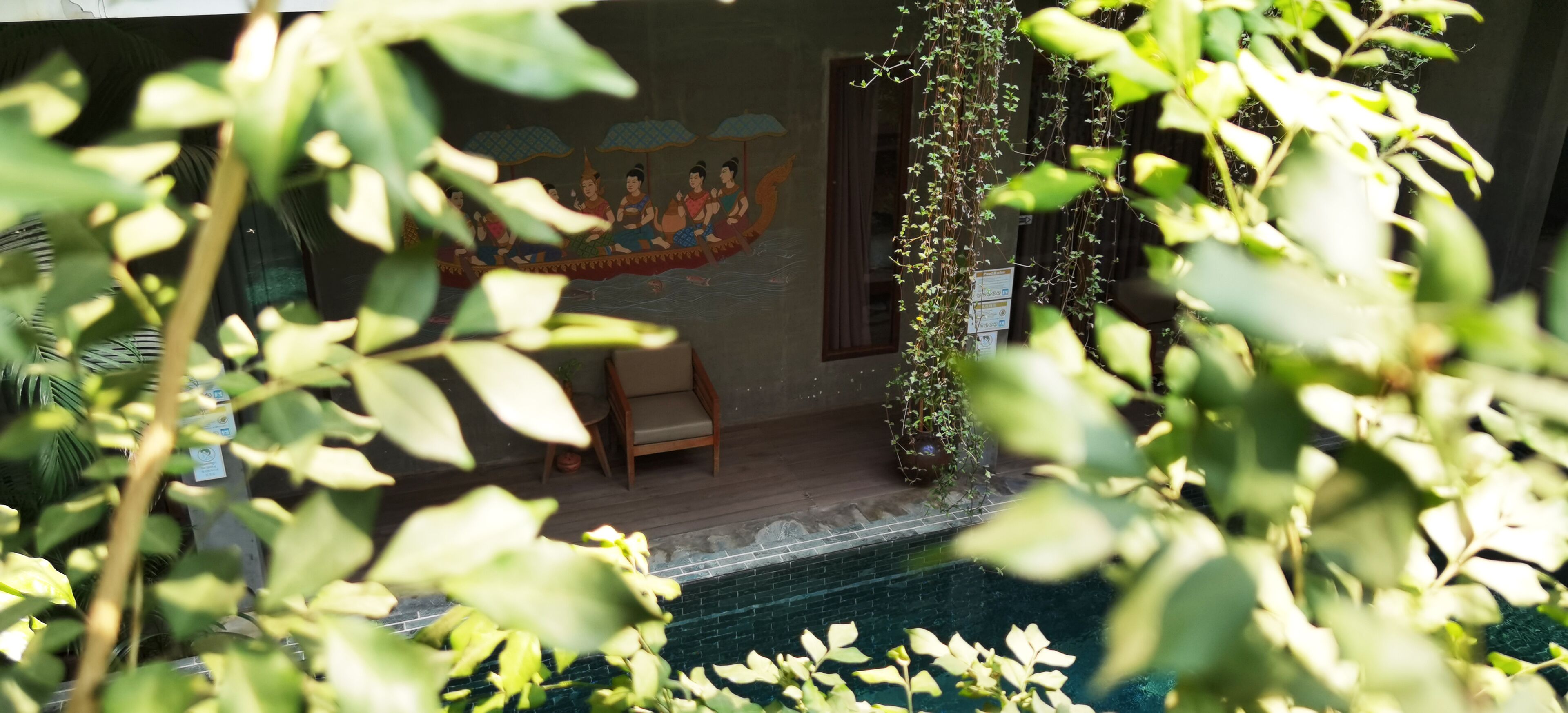 Un mural tailandés tradicional, una terraza de madera con sillas y una piscina.