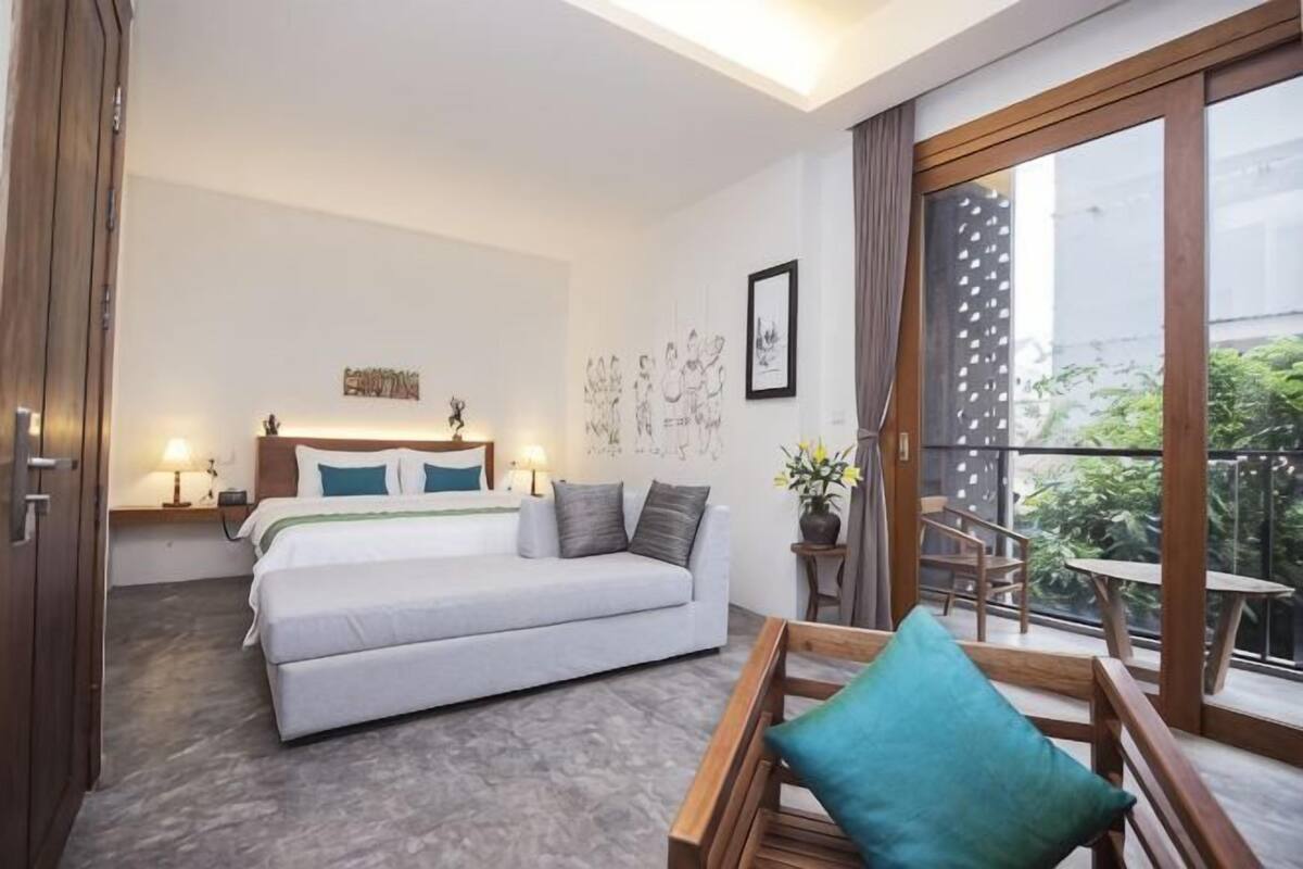 Une chambre avec un lit, des tables de chevet, un canapé, un balcon avec une table et des chaises, et une vue sur la verdure environnante.