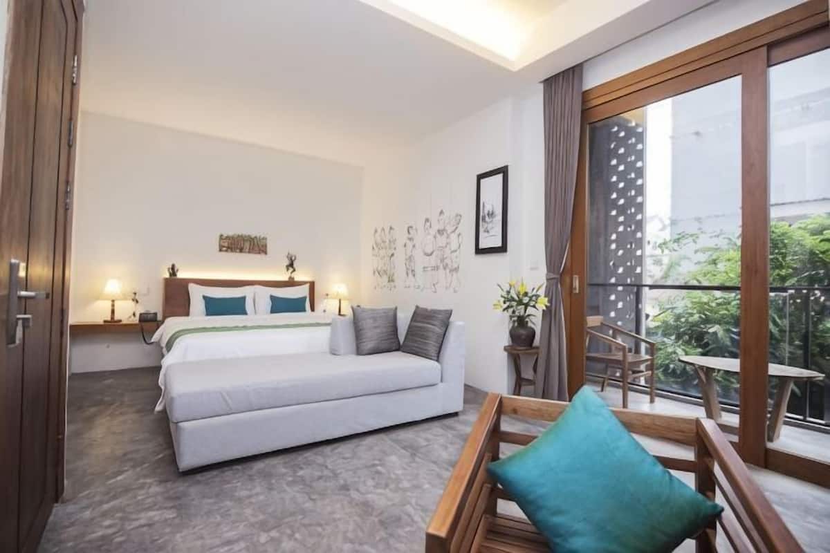 Quarto com cama, mesas de cabeceira, um sofá, uma varanda com mesa e cadeiras, e vista para o verde lá fora.