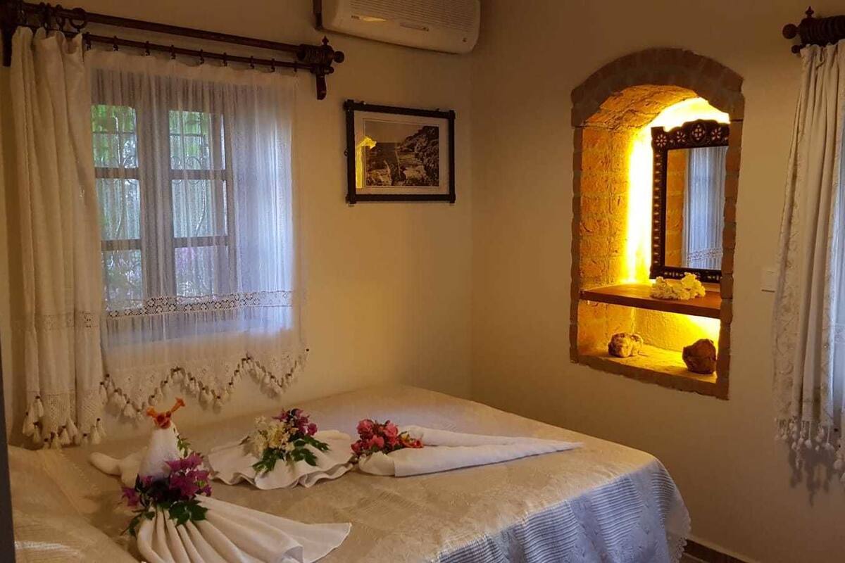 Una cama bien hecha con edredón blanco y adornos florales, un pequeño espejo arriba y una ventana con arco de piedra.