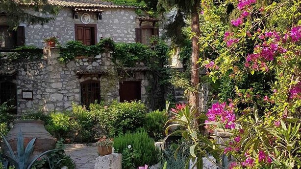 A stone building with a tiled roof, surrounded by lush greenery and flowering plants.