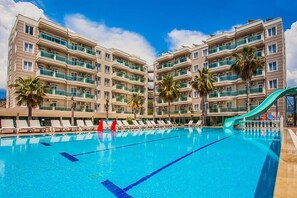 A swimming pool with lounge chairs and palm trees, surrounded by multi-story buildings with balconies. - Grand Akca Hotel (Gazipasa)