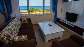 A living room with a sofa, a coffee table, and a TV. There is a large window with a view of the ocean and buildings. - Grand Akca Hotel (Gazipasa)