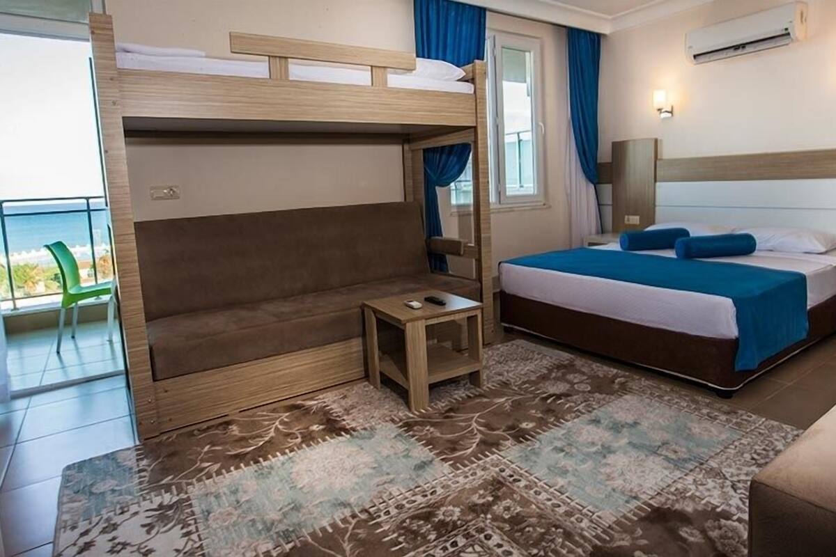 A room with a bunk bed, a sofa, a small table, and a large bed with blue bedding. There is a window with curtains, a wall-mounted air conditioner, and a green chair outside the window.