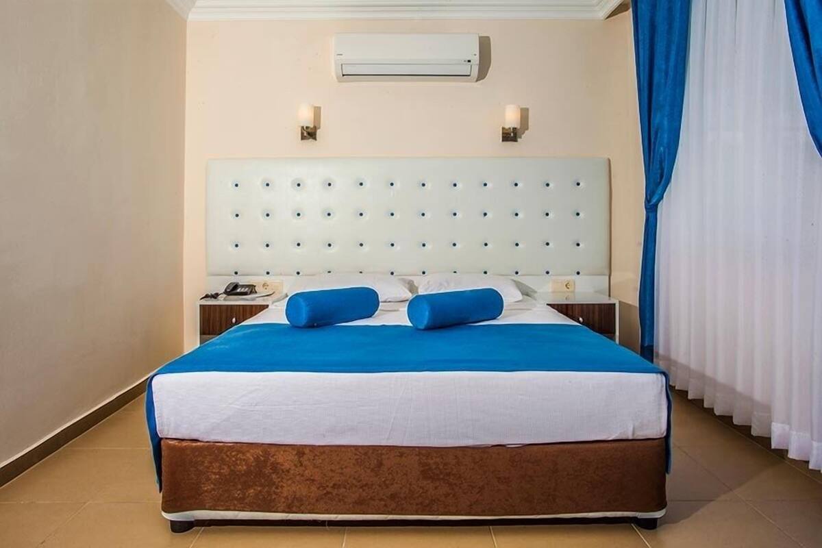 A hotel room with a large bed, a white headboard, two bedside lamps, a telephone, and a window with curtains.