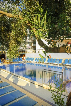 A swimming pool with blue lounge chairs and a lifebuoy, surrounded by trees and a building with balconies. - Mutlu Apart Otel (Didim)