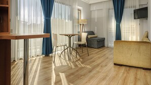A hotel room with a wooden floor, a sofa, a dining table with chairs, and a TV mounted on the wall. - Mutlu Apart Otel (Didim)