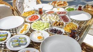 An outdoor dining area with wicker furniture, a variety of food and drinks, and a view of the hotel building in the background. - Mutlu Apart Otel (Didim)