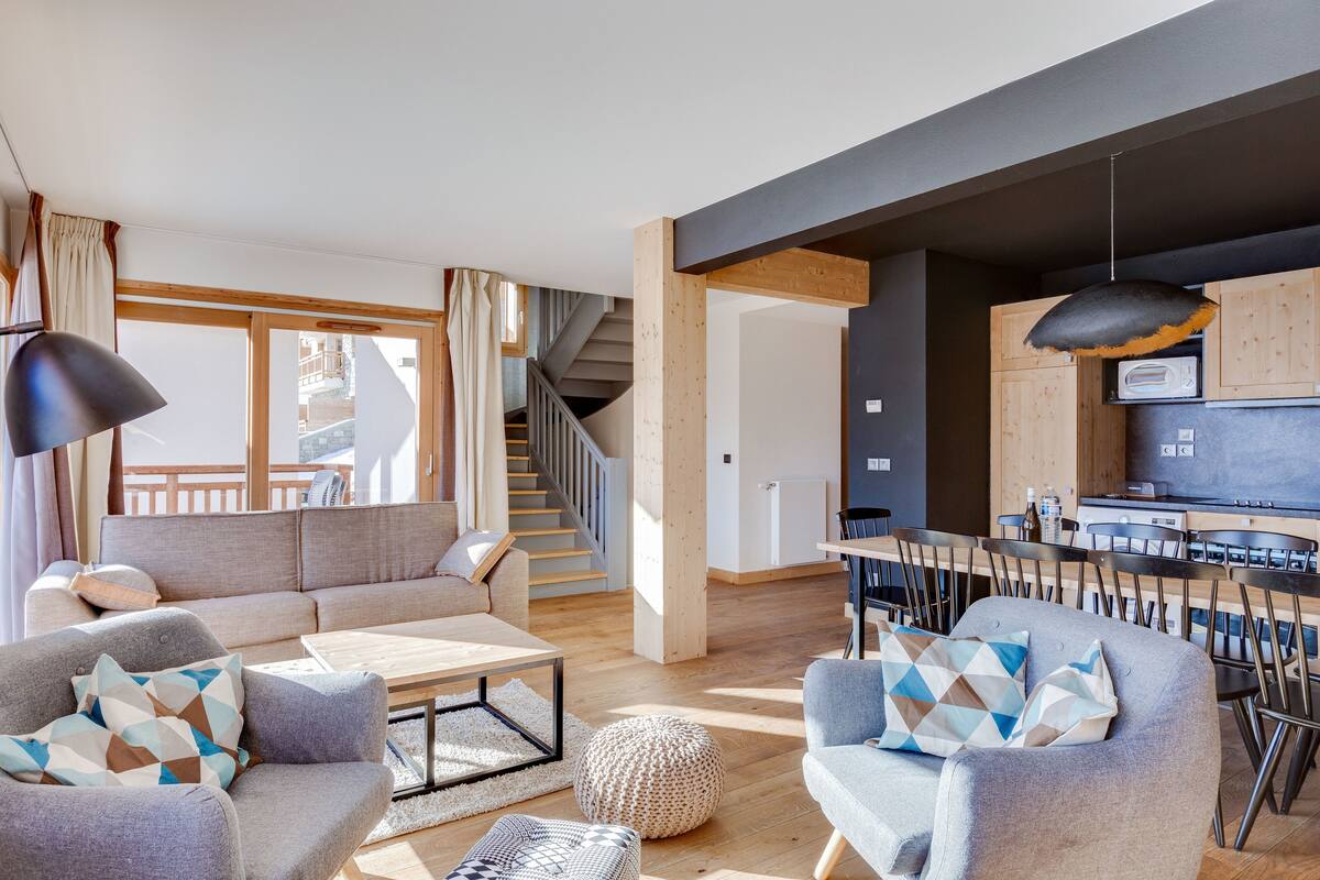 Un salon moderne avec un canapé, des fauteuils et une table basse. Un escalier mène à un espace cuisine doté de meubles en bois et d’une table à manger.