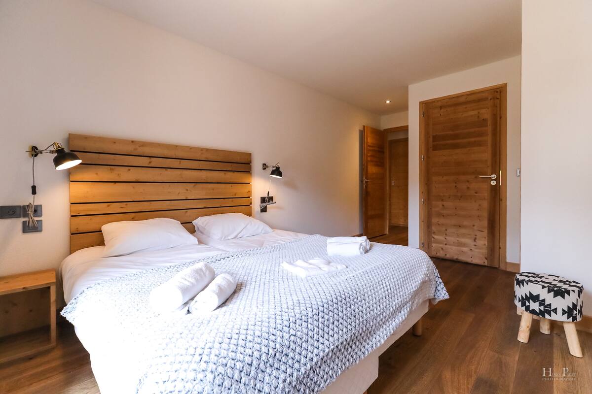Un lit bien fait, avec du linge de lit et des serviettes blanches, une tête de lit en bois, des lampes de chevet et un petit tabouret en bois.