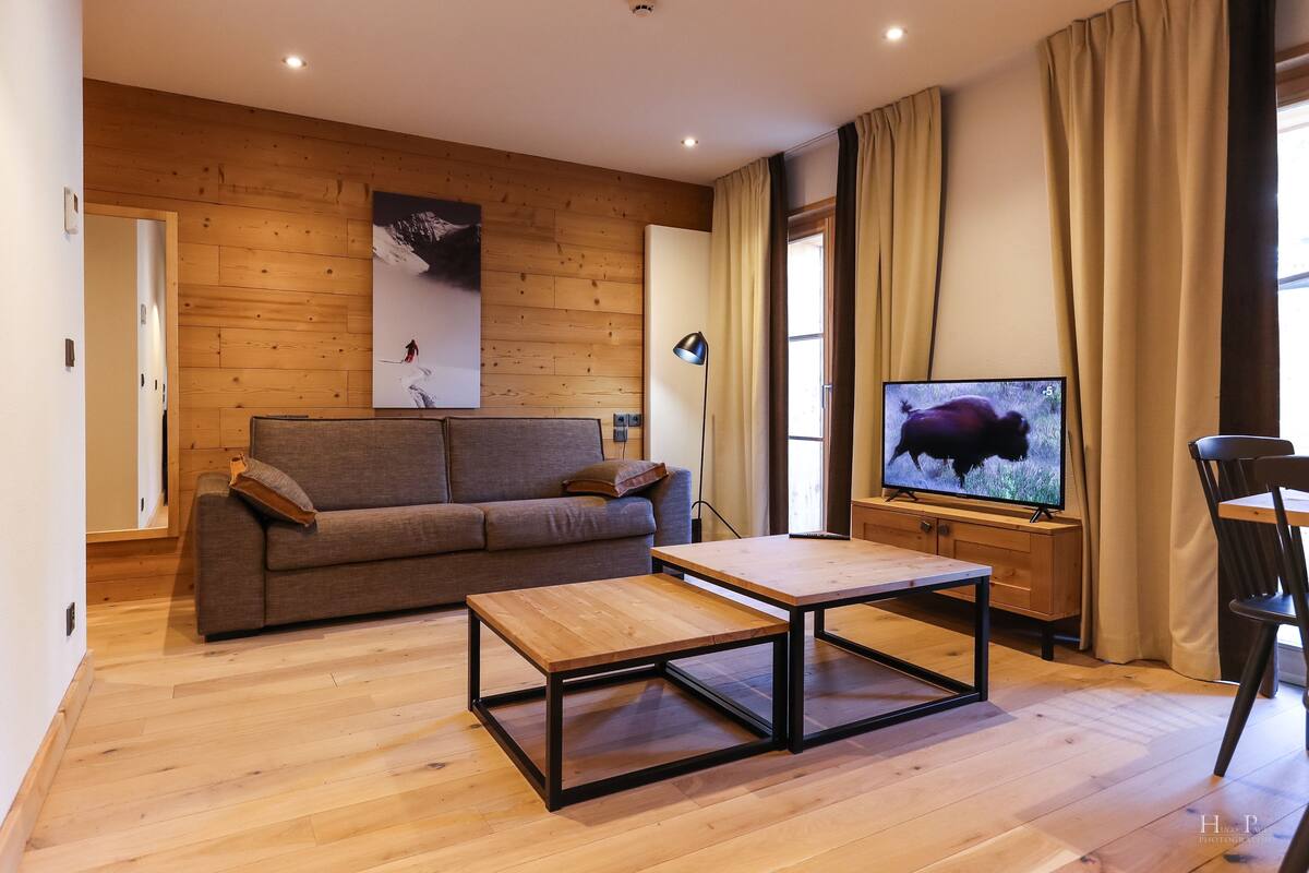 Un salon moderne avec un plancher en bois, un canapé, une table basse, un téléviseur et une fenêtre avec des rideaux.