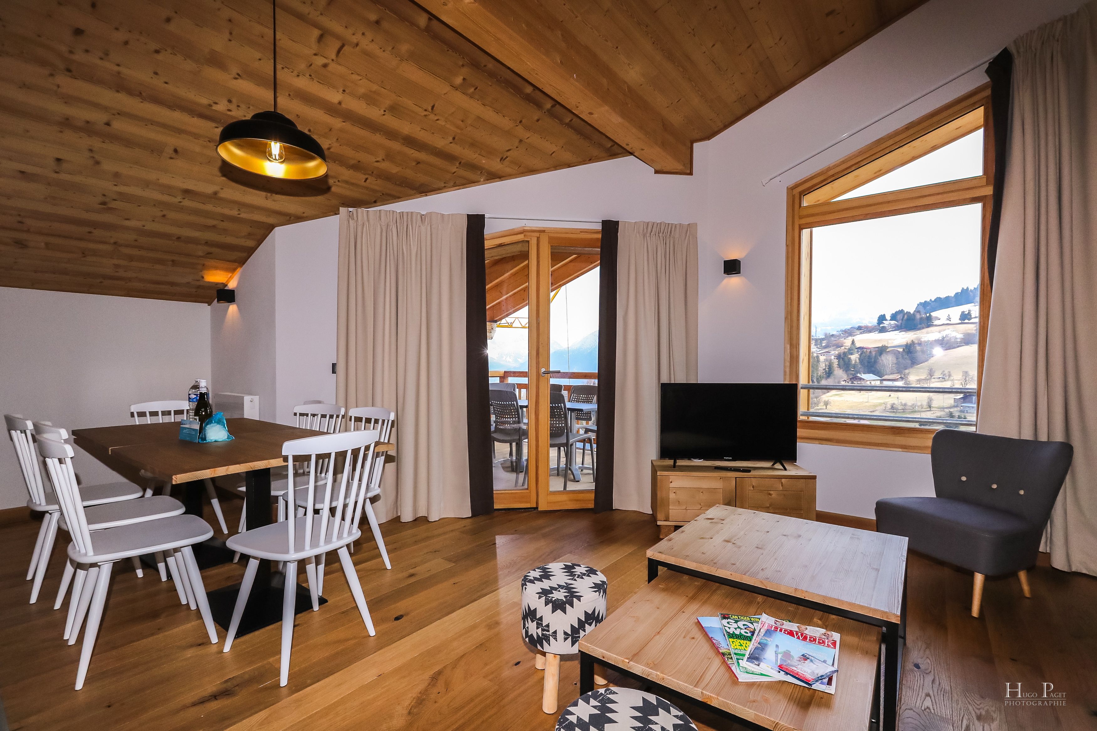 A wooden ceiling, a dining table with chairs, a sofa, a TV, and a view of a snowy landscape through a large window.