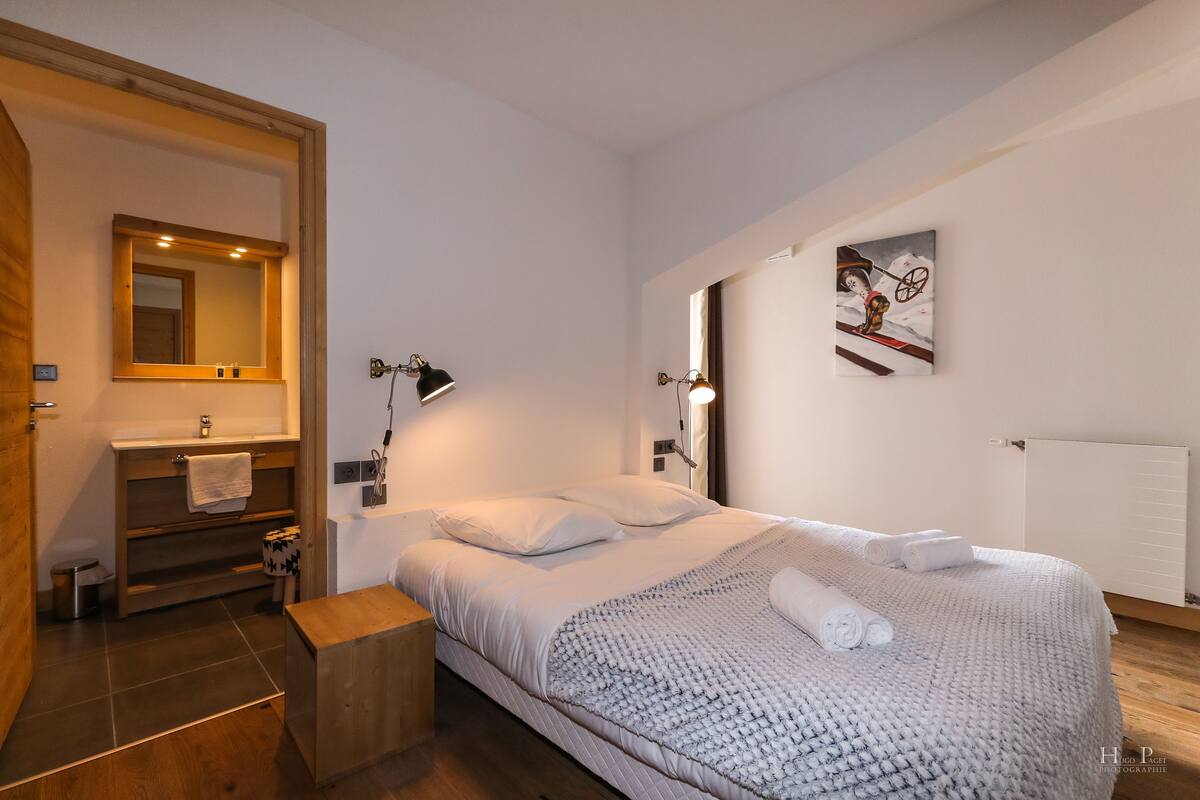 Une chambre à coucher comprenant un lit, une table de chevet, une commode, un miroir, une lampe fixée au mur et un tableau.