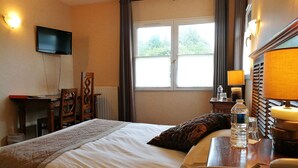 A hotel room with a bed, a desk with a chair, a television, a window with curtains, a lamp, and a telephone.