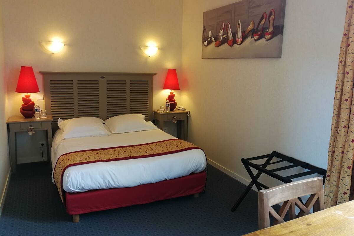 Een hotelkamer met een bed, twee nachtkastjes met lampen, een kleine tafel, een stoel en een schilderij aan de muur.