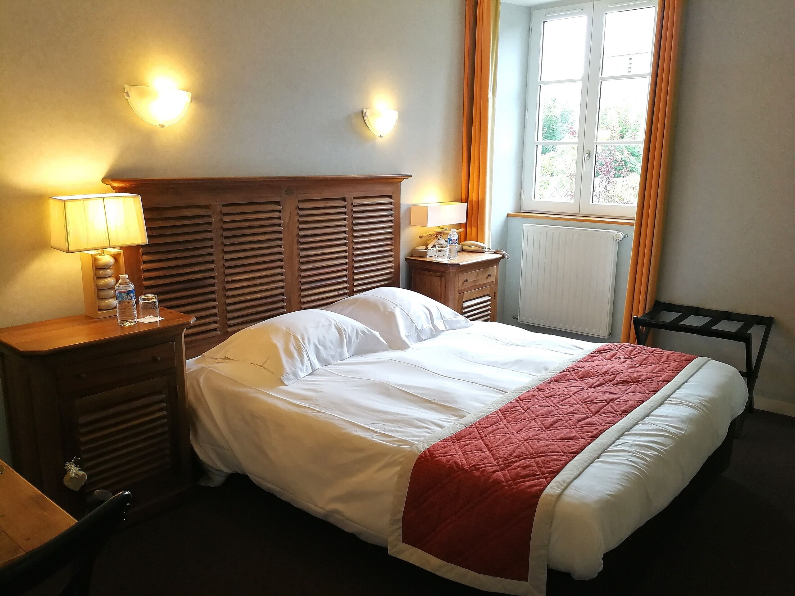 Et hotelværelse med en seng, to sengeborde med lamper, en træsenggegærde, et natbord med en telefon og en flaske, samt et vindue med gardiner.