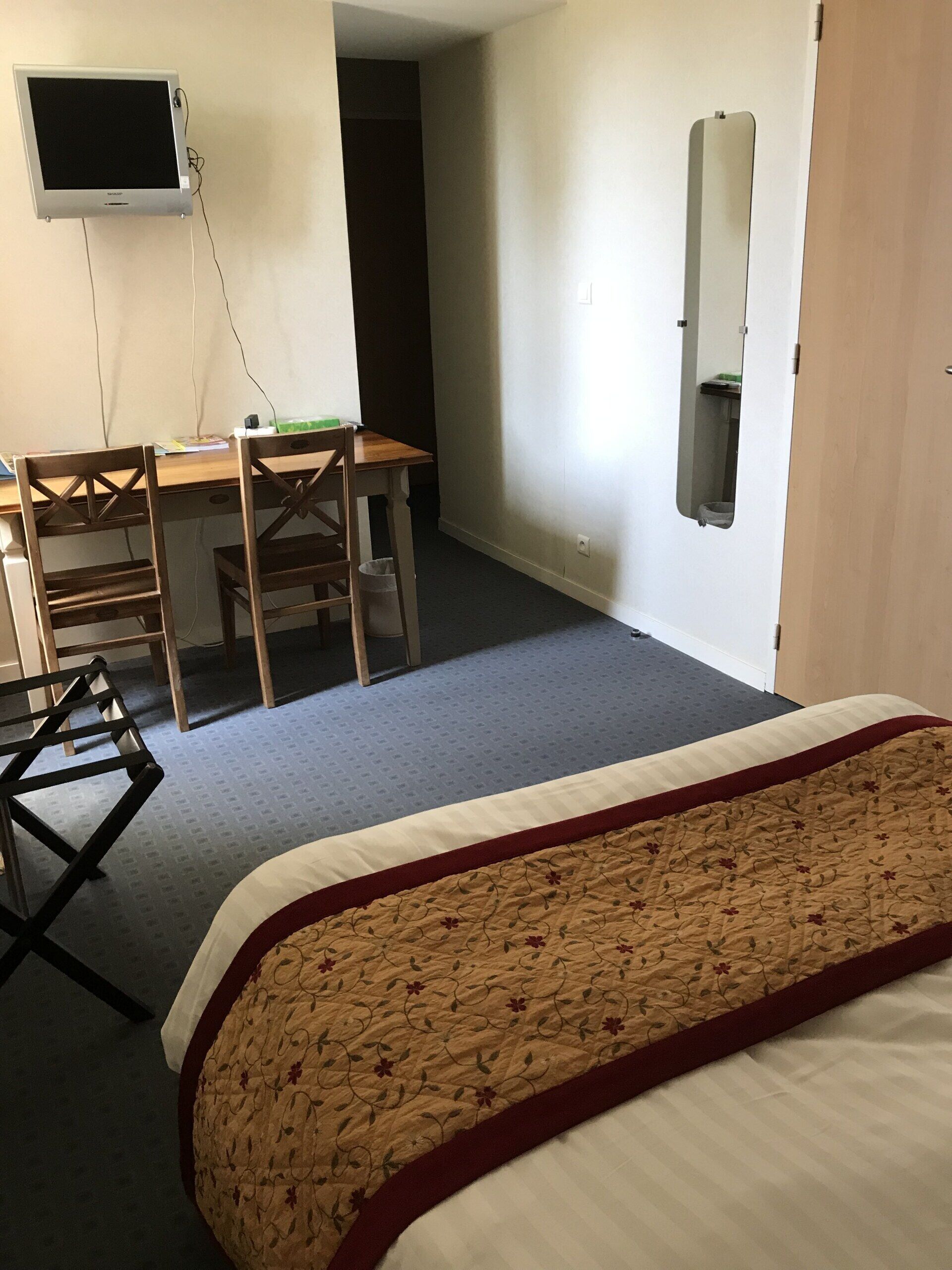 A hotel room with a bed, a desk with a chair, a television mounted on the wall, and a mirror.