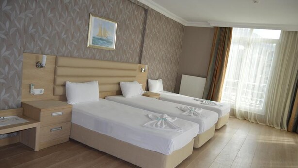 A hotel room with two beds, a wooden headboard, a bedside table, and a window with curtains. - Hotel Kale Palace (Gökçeada)