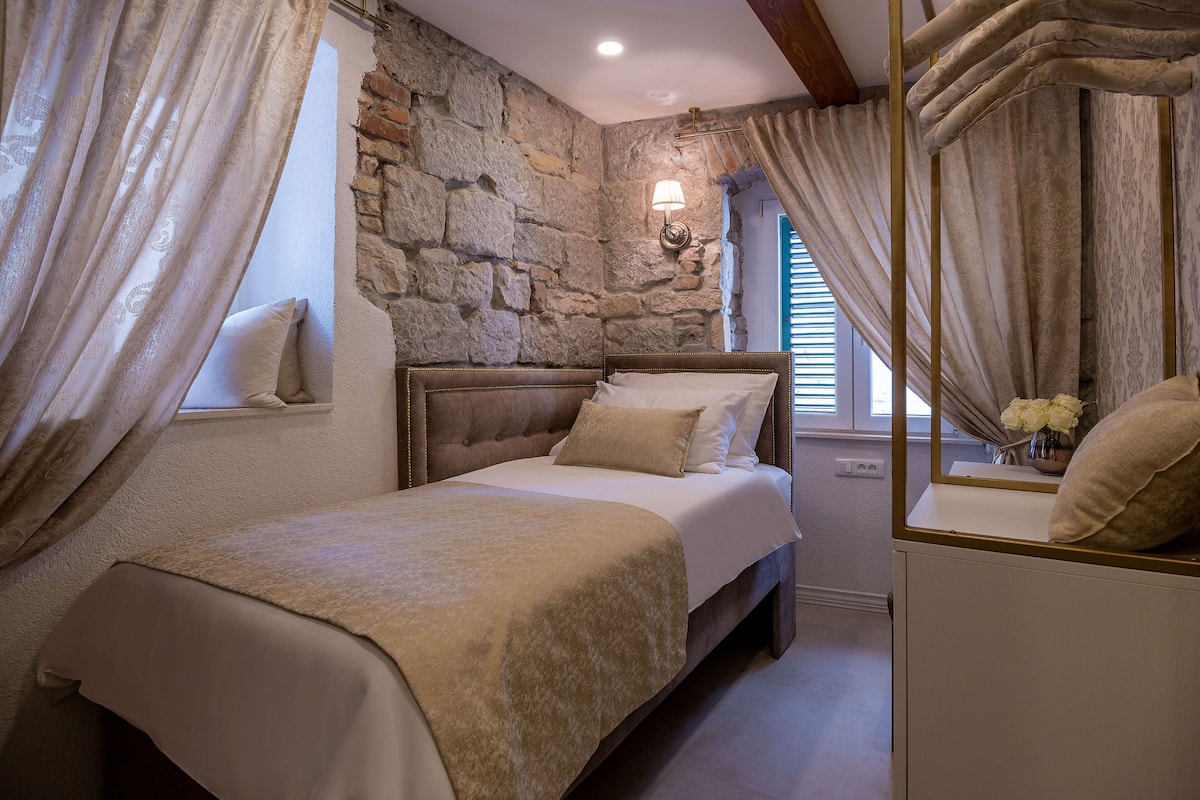 A bedroom with a stone wall, a bed with a beige bedspread, and a nightstand with a vase of flowers.
