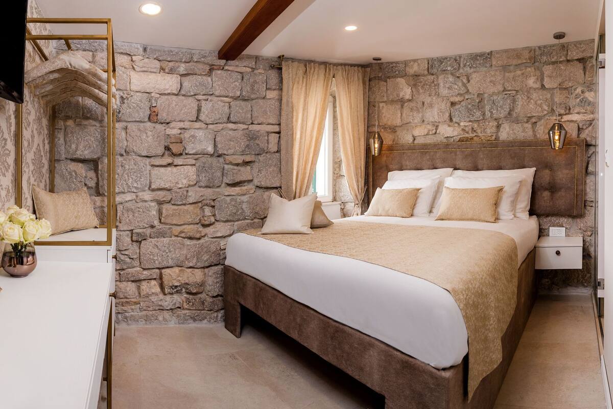 A bedroom with a stone wall, a bed with multiple pillows, a bedside table, and a mirror.