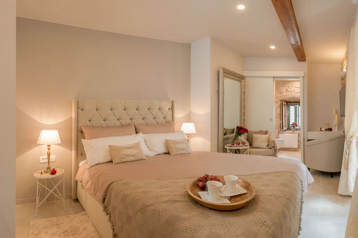 A spacious bedroom with a large bed, bedside lamps, a wooden tray with cups and grapes, and a view into the bathroom.