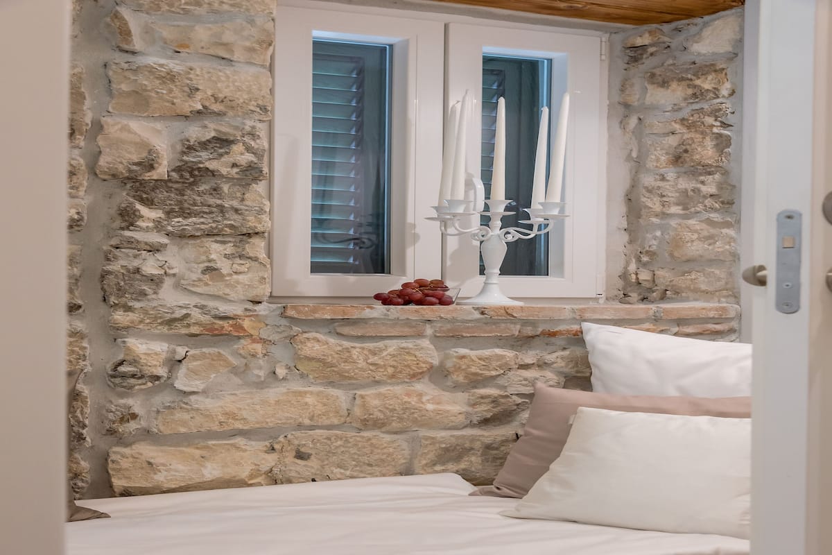 A bedroom with a stone wall, a window with shutters, a candelabra, and a bed with white linens.
