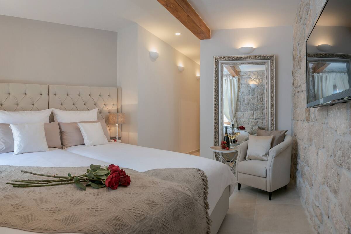 A bedroom with a large bed, a bedside table with a lamp, a chair, a mirror, and a bouquet of flowers.