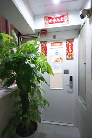 Un pasillo estrecho con una planta en maceta a la izquierda, una puerta con un cartel rojo encima y una cámara de seguridad montada en la pared.