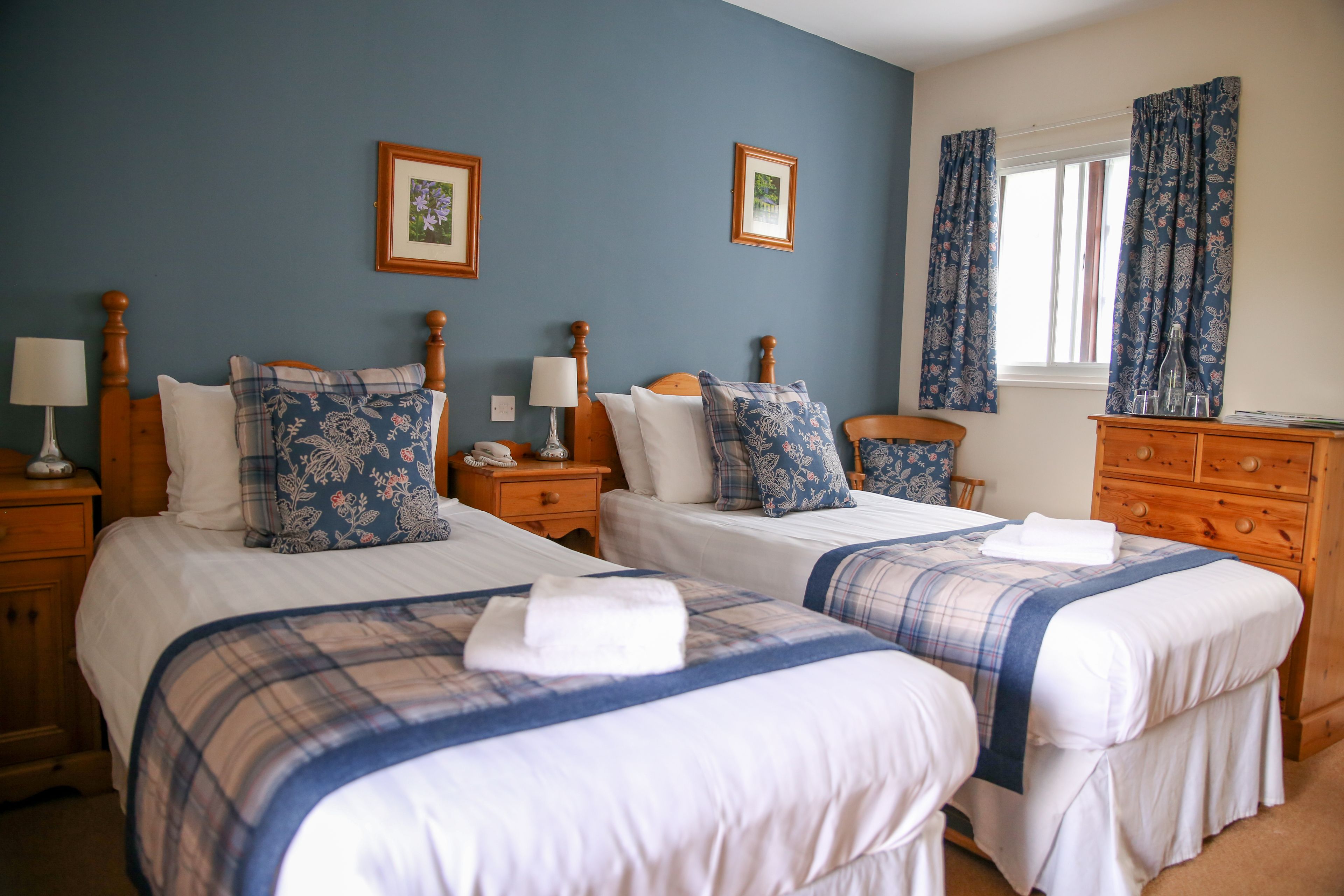 Quarto de hotel com duas camas, cada uma com colcha xadrez azul e branca, cabeceiras de madeira e almofadas a combinar. Há duas mesas de cabeceira com abajures, uma cômoda de madeira e uma janela com cortinas.