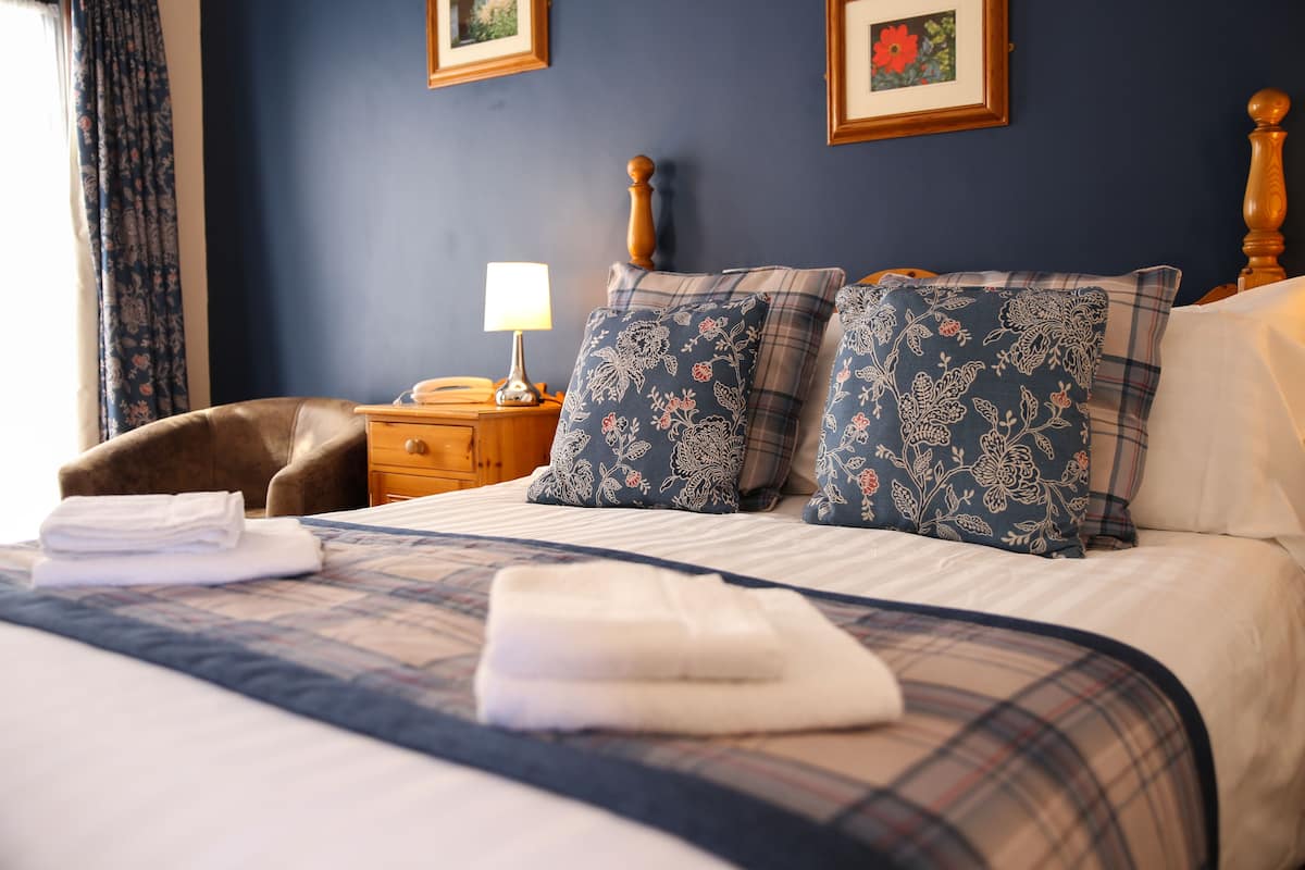 A neatly made bed with a plaid bedspread, two pillows with floral patterns, a bedside lamp, and a wooden nightstand.