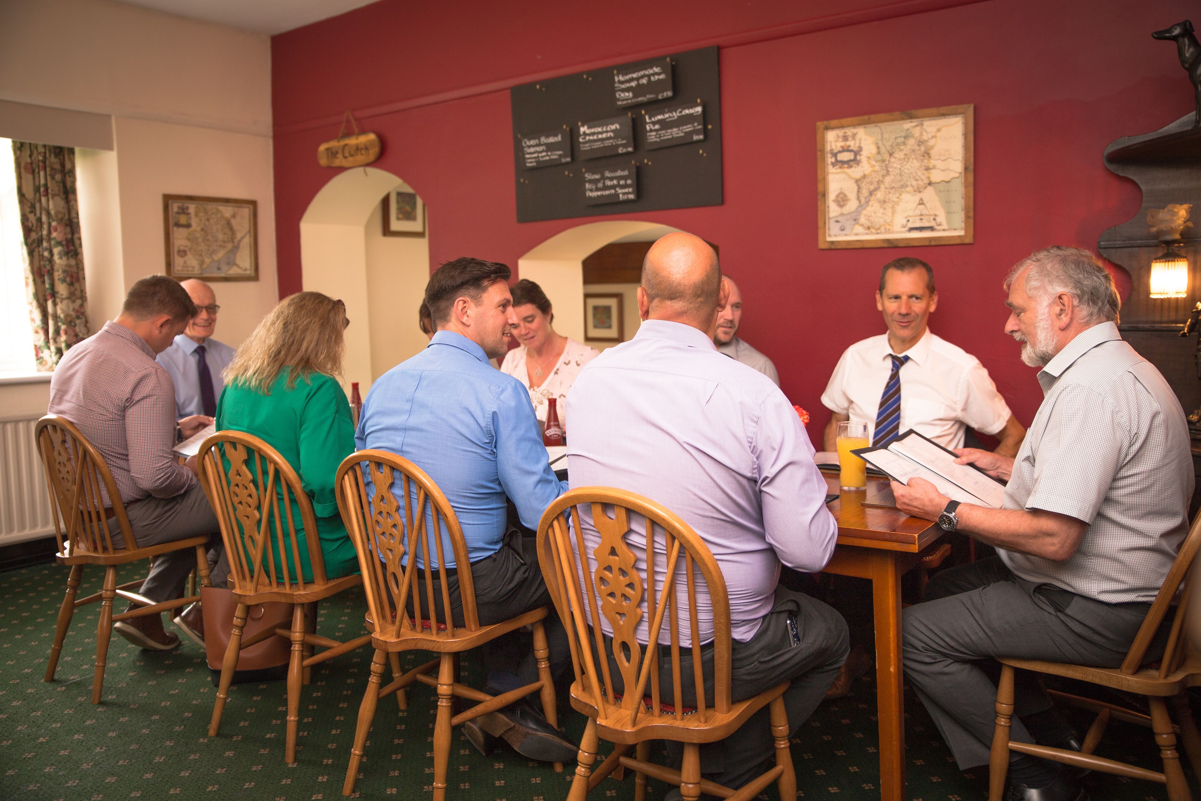 A group of people seated around a wooden table in a room with red walls, a map on the wall, and a menu board.