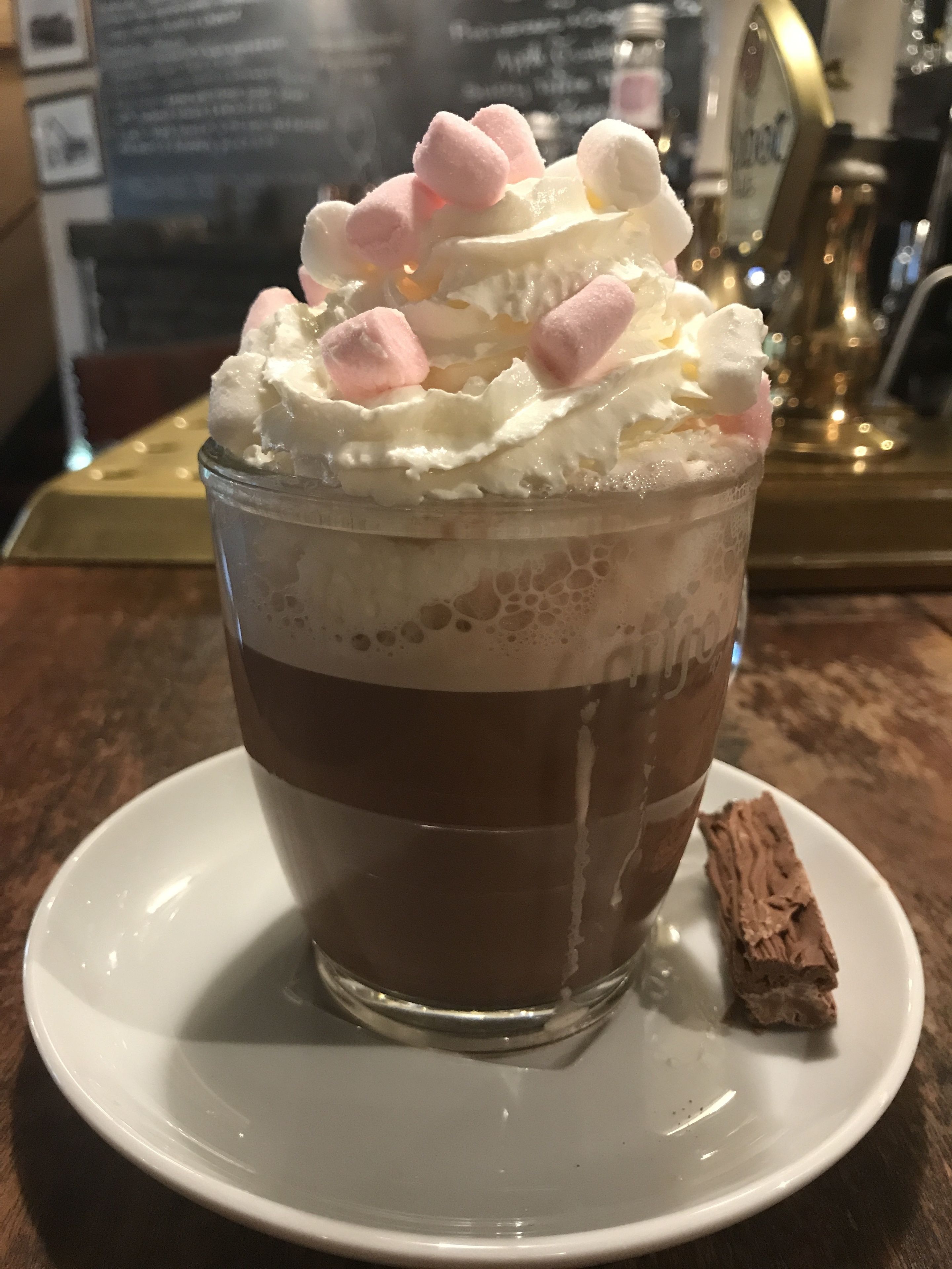 A layered dessert in a glass cup with whipped cream and marshmallows on top, served on a white plate with a chocolate piece beside it.