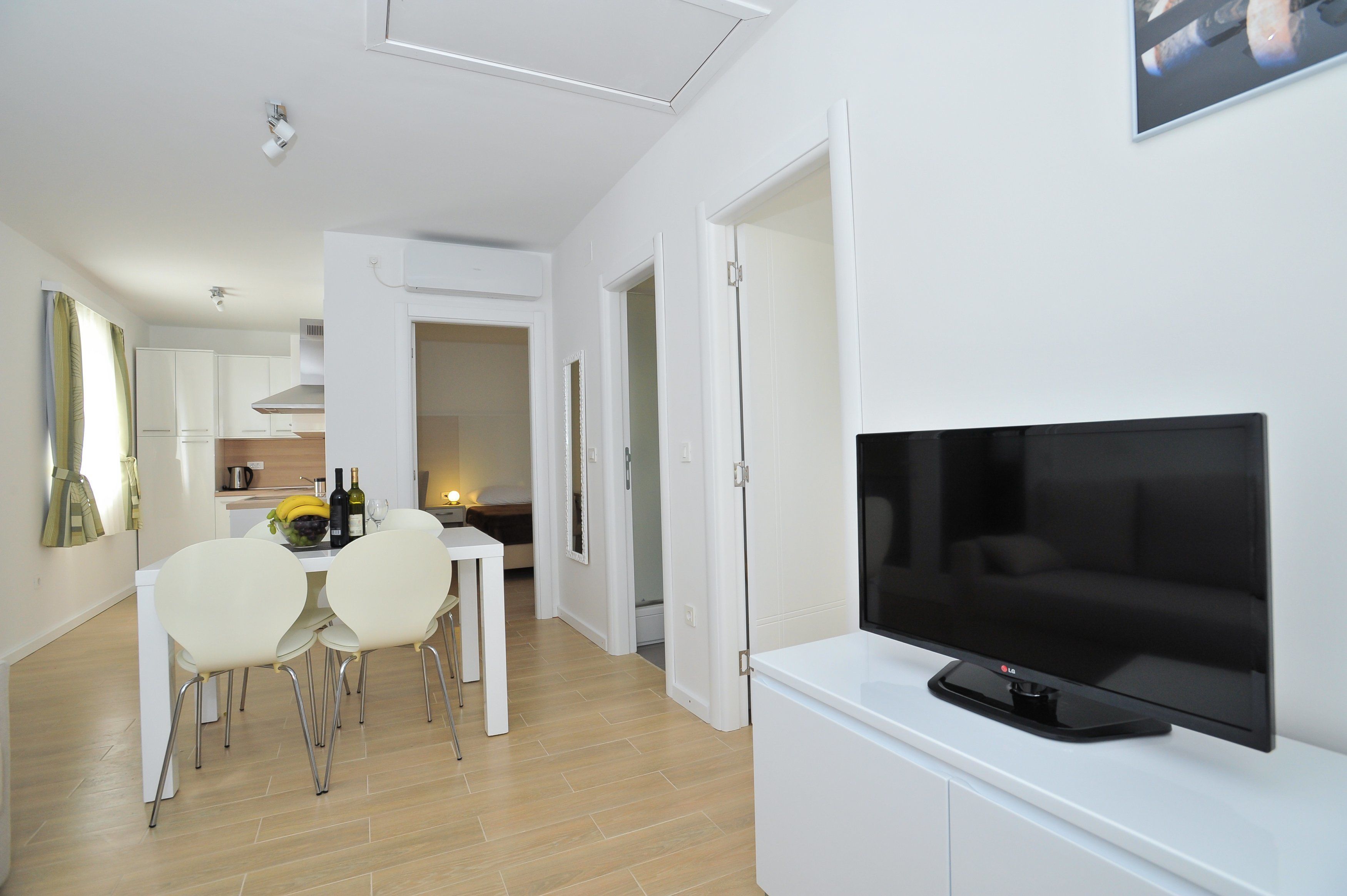Un soggiorno moderno con una televisione a schermo piatto su un mobile bianco, una zona pranzo con sedie e tavolo bianchi e una cucina sullo sfondo.