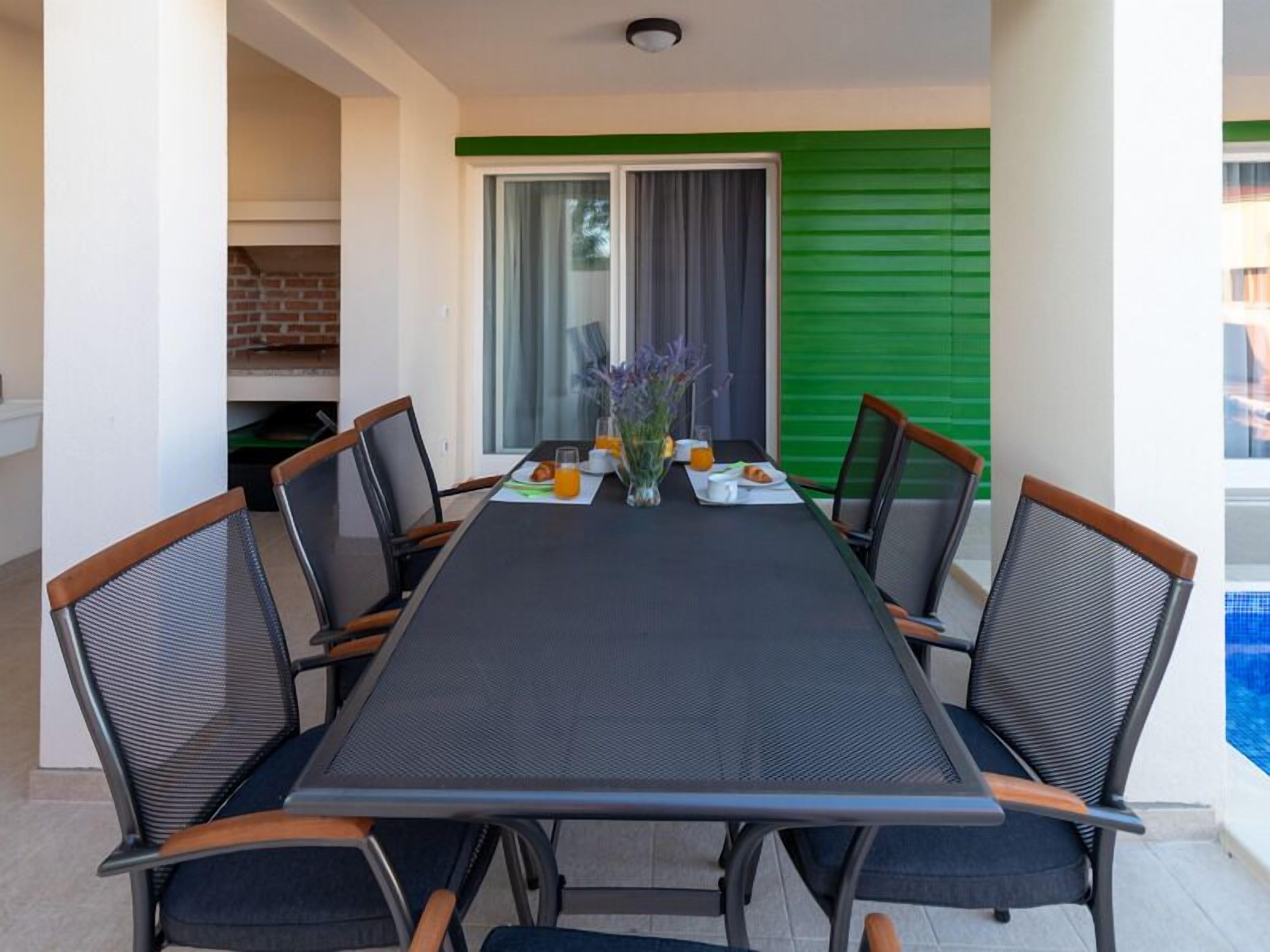 A dining area with a table set for six, surrounded by chairs with mesh backs and wooden armrests.
