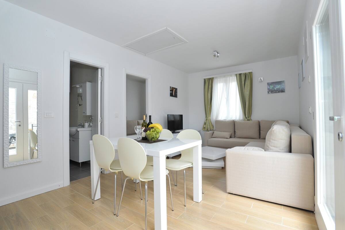 Ein modernes Wohnzimmer mit einem weißen Sofa, einem Esstisch mit Stühlen und einer Küche im Hintergrund.