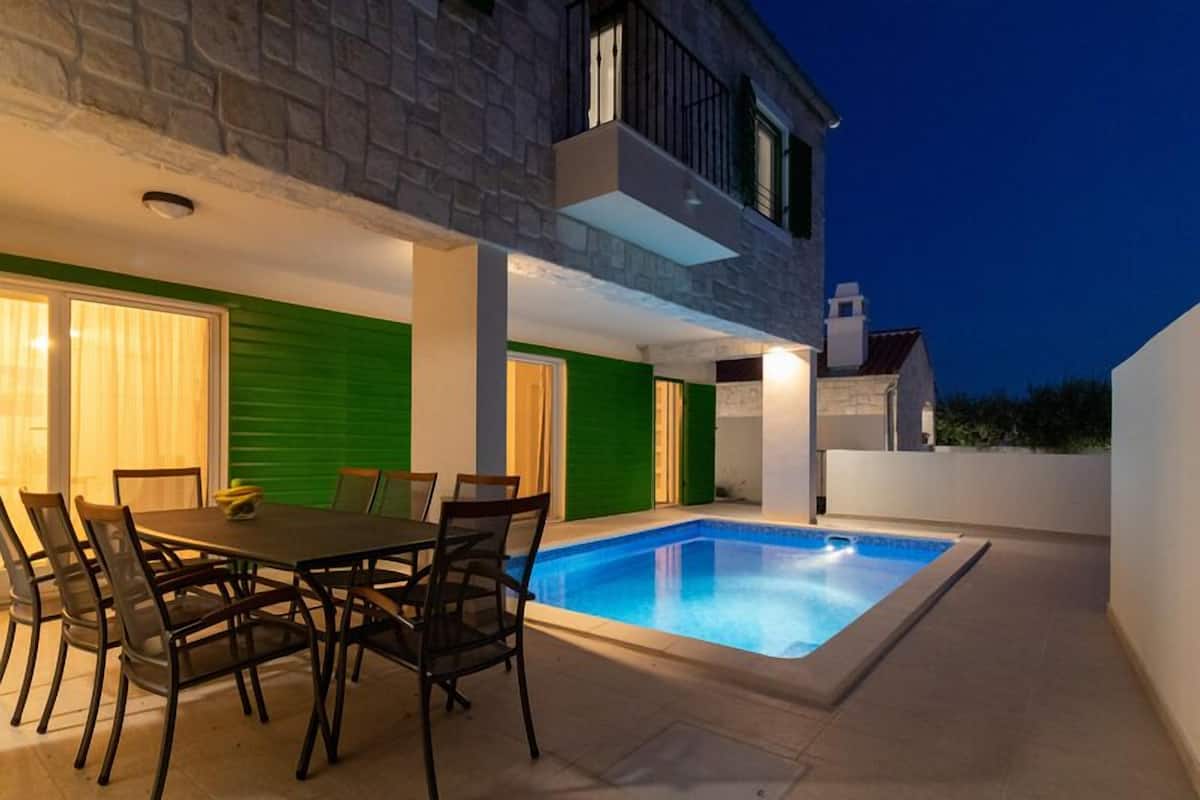 Ein modernes Haus mit Swimmingpool, einem Essbereich inklusive Tisch und Stühlen, und einer türfarbigen, grün schattierten Tür.