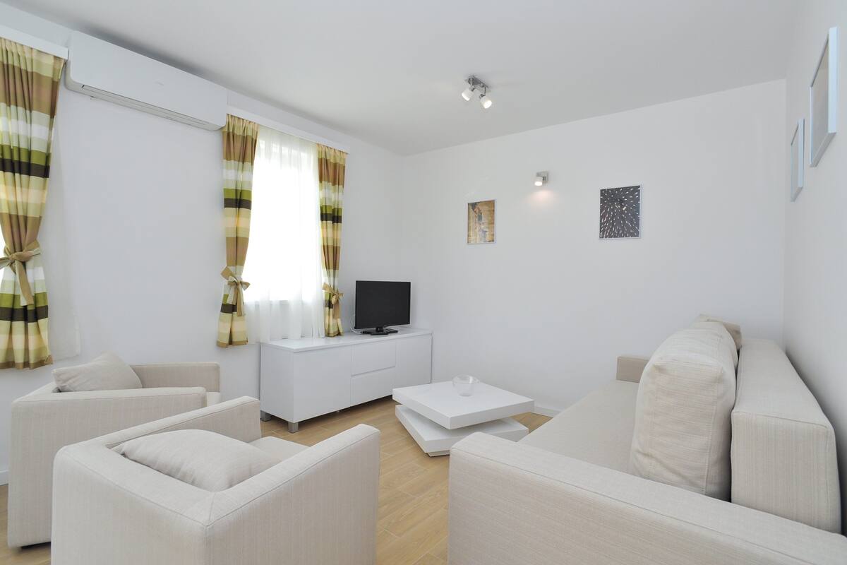 Ein modernes Wohnzimmer mit einer Couch, Sesseln, einem Couchtisch, einem Fernseher und Vorhängen.