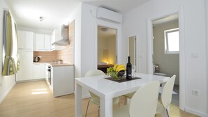 A modern kitchen with white cabinets, a built-in oven, and a dining area with a white table and chairs. - Apartments Corte (Privlaka)