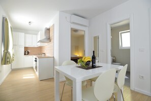A modern kitchen with white cabinets, a built-in oven, and a dining area with a white table and chairs. - Apartments Corte (Privlaka)