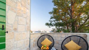 A stone wall with a green door, two black wicker chairs with yellow cushions, a small round table with fruit and cups, and a view of trees and buildings. - Apartments Corte (Privlaka)