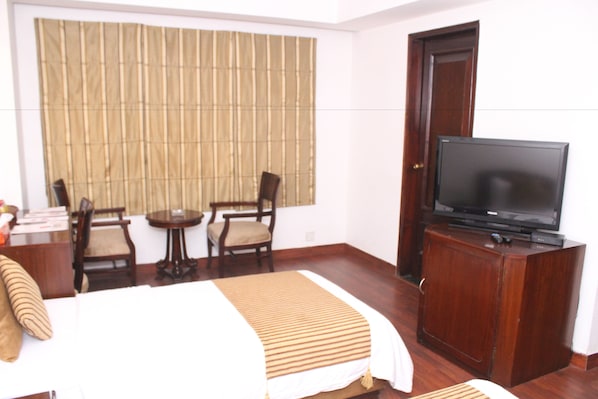 A hotel room with a bed, a television on a wooden cabinet, a small table with chairs, and a window with vertical blinds.
