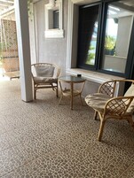 Una habitación con suelo de baldosas, dos sillas de mimbre, una mesita redonda y una ventana con vistas a zonas verdes.