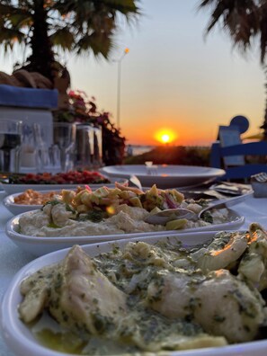 A table set for a meal with various dishes, a sunset in the background, and a palm tree. - Kale Motel (Gökçeada)